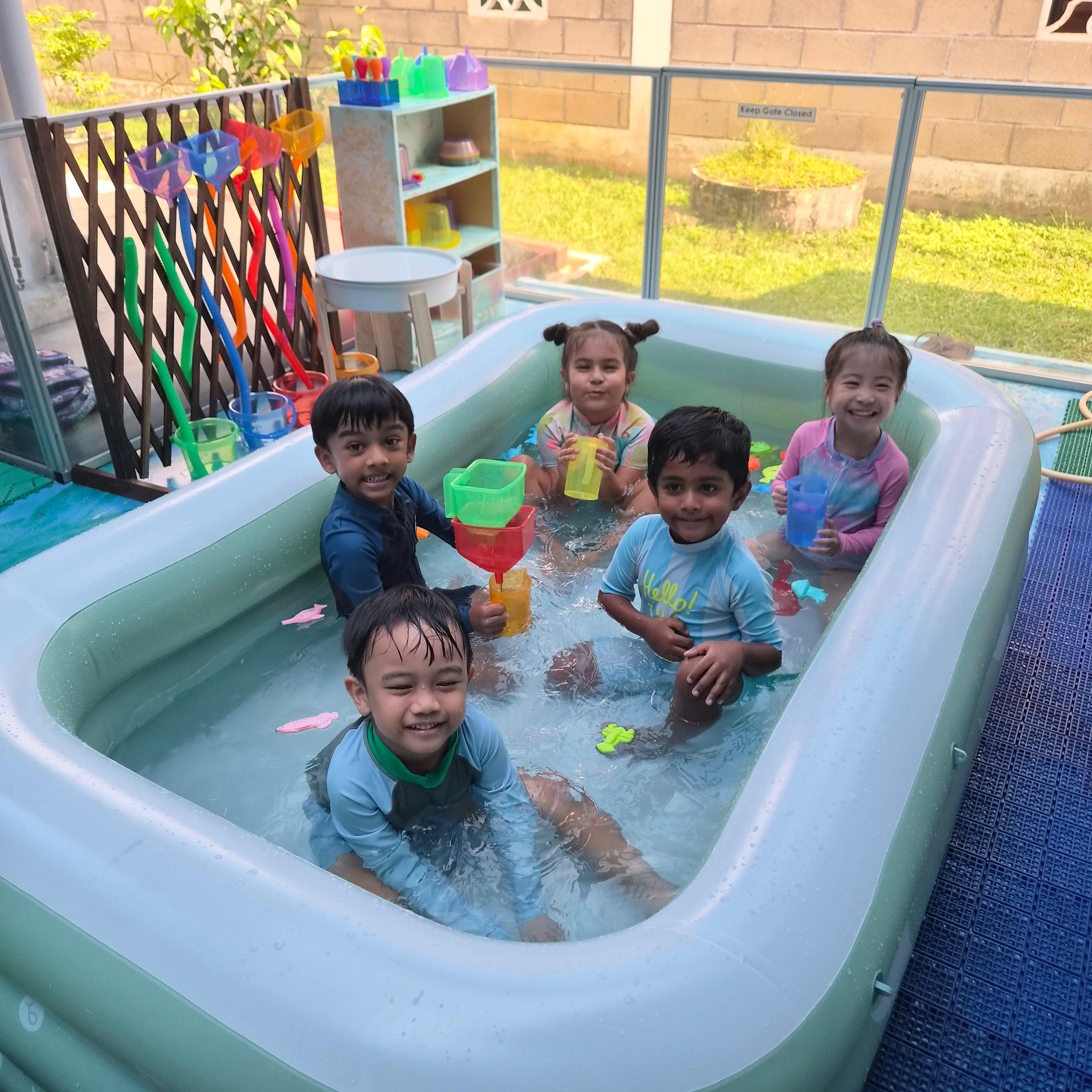 Five children splashing and playing with water toys in an outdoor paddling pool at Genius Gems Childcare Centre
